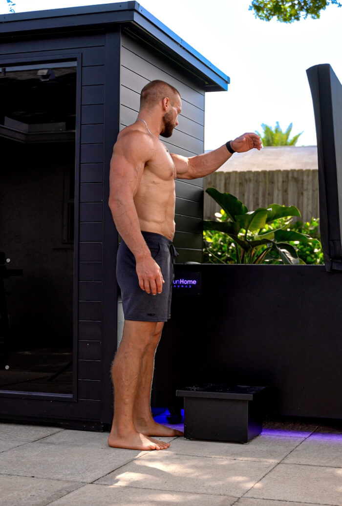 Muscular man standing shirtless next to a modern black outdoor sauna and cold plunge tub on a patio.