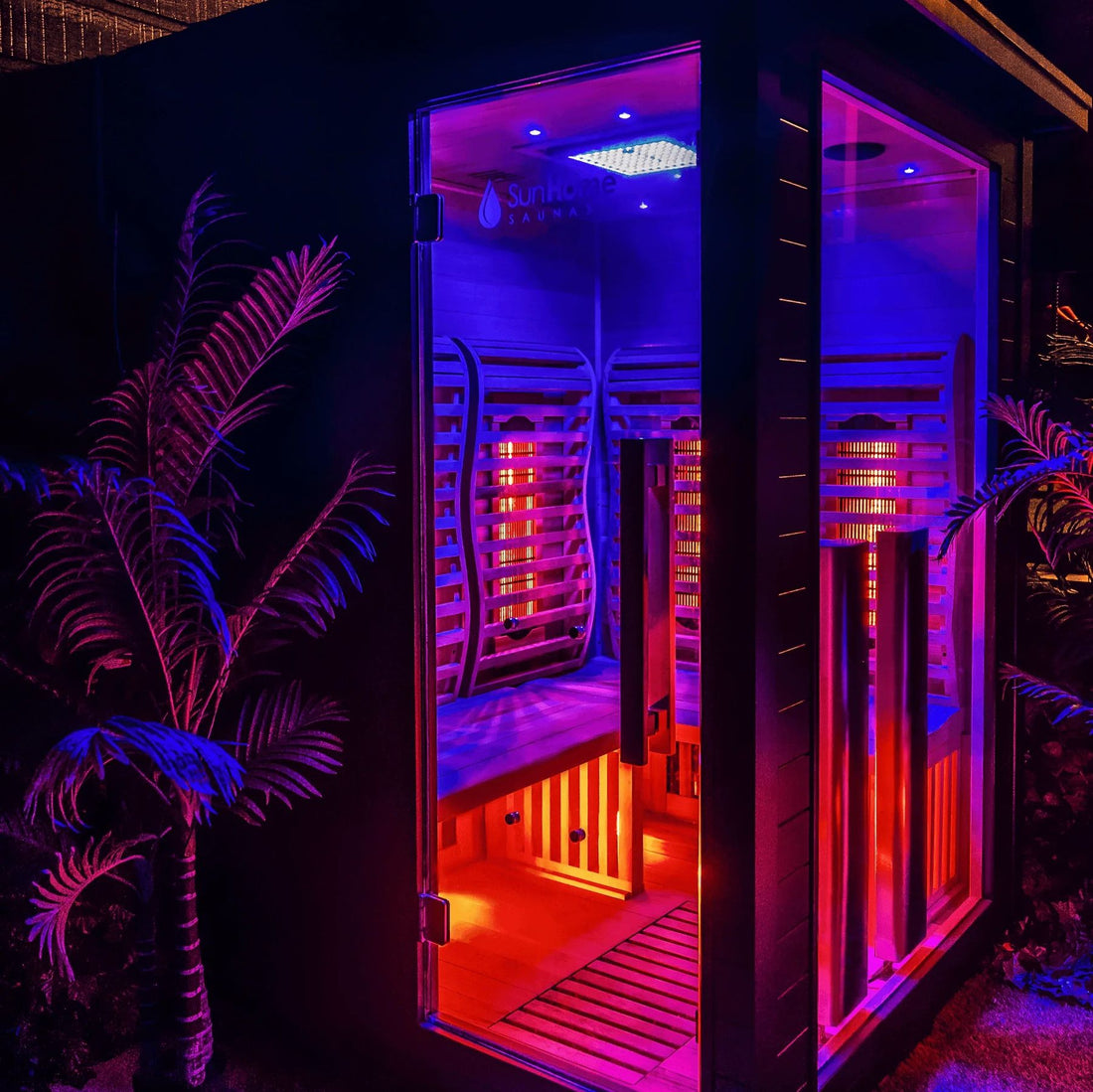 Sun Home Luminar 2-person outdoor infrared sauna at night with its glass door open, glowing with blue and red chromotherapy lighting and infrared heaters