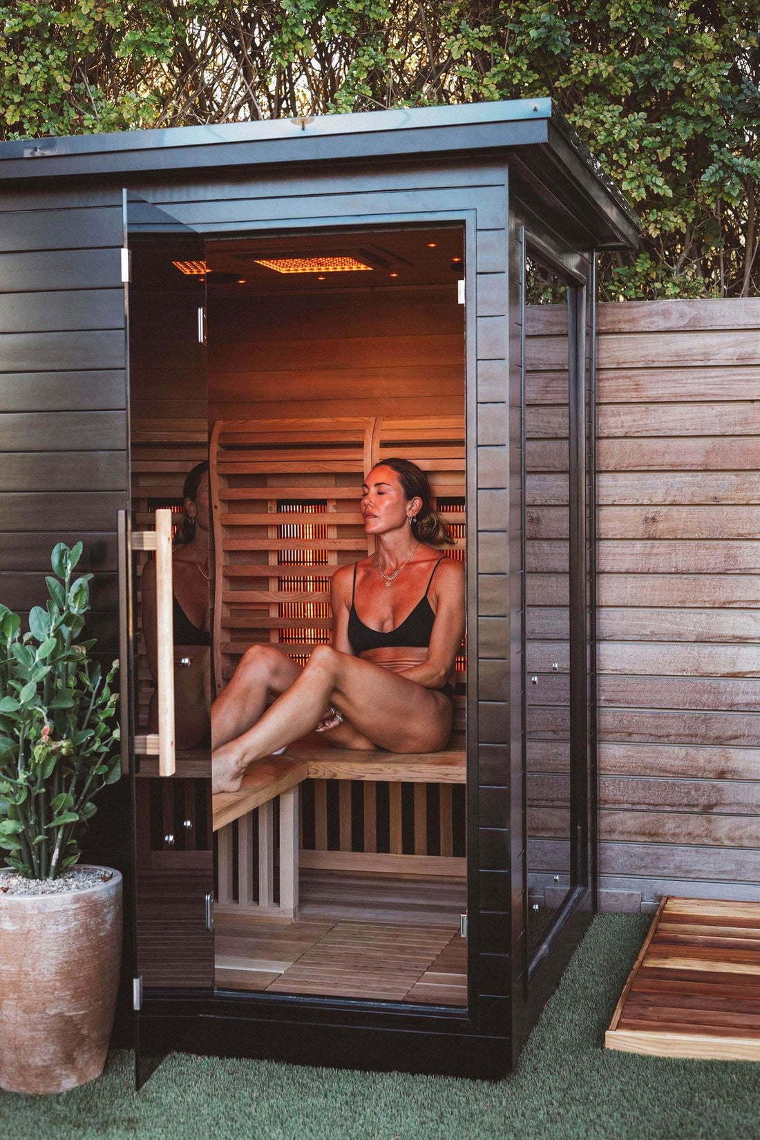 Woman sitting inside the Sun Home Luminar 2-person infrared sauna with the glass door open, surrounded by far-infrared heater panels