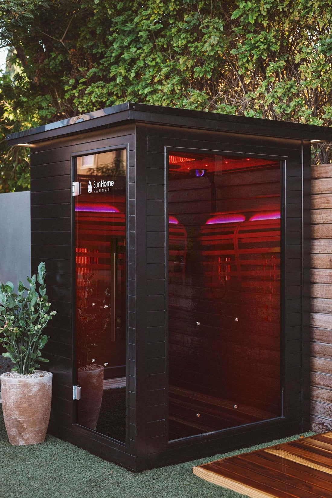 Sun Home Luminar 2-person outdoor infrared sauna on a patio with red and purple chromotherapy lighting glowing through its panoramic black glass